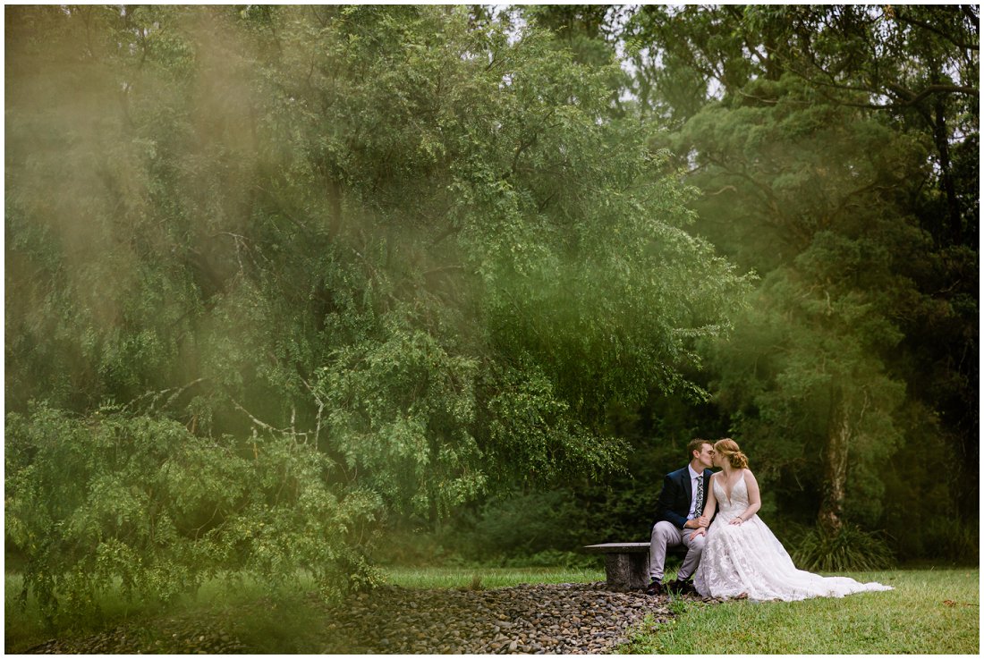 bride and groom sitting on park bench in the rain